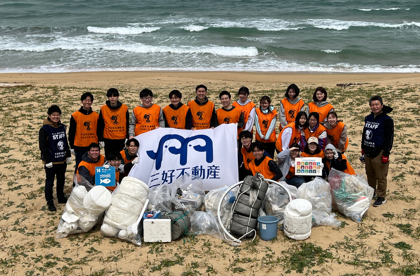 ビーチクリーン実施時、拾った大変多くのゴミを前に海辺で20名ほどがいる集合写真